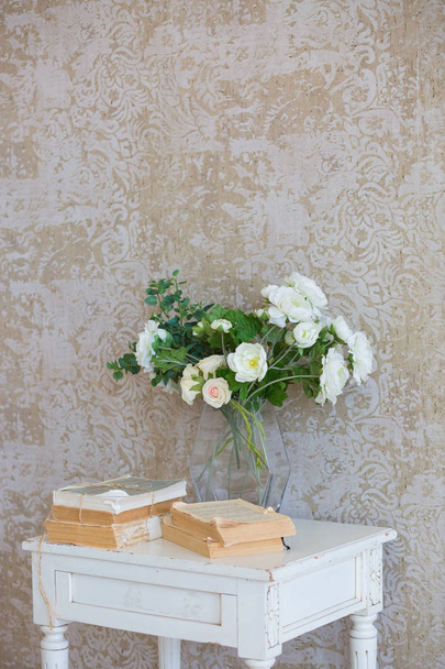 White bedside table against a beige wall. There are old books on the table and a vase of white flowers. Near the pedestal is a white floor candlestick lantern. An empty white frame hangs on the wall. Classic interior with white wooden floor. - Foto, imagen