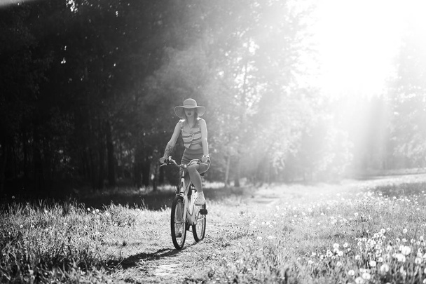 Funny girl driving bicycle outdoor. Sunny summer lifestyle concept. Woman in dress and hat in Field with dandelions. Female ride in park. Light effect for text. Copyspace for design. BW photo. - Foto, afbeelding