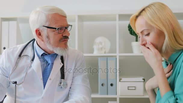 Coughing sick female patient complaining to caucasian midle aged doctor in glasses sitting in office about her throat. - Materiał filmowy, wideo