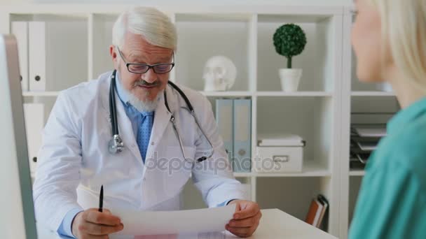 Caucasian midle aged male doctor in glasses sitting in office, smiling to young female patient, reading something from sheet of paper. Indoor. - Felvétel, videó
