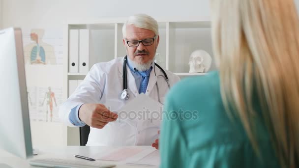 Caucasian serious midle aged doctor in white robe with glasses and beard sitting in his office explaining something using sheet of paper. Indoor. - Video