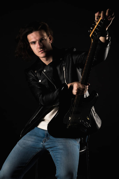 Hudba a kreativity. Pohledný mladý muž v tričko, bundu a džíny, s elektrickou kytarou, na černém pozadí, samostatný. Vertikální rám - Fotografie, Obrázek