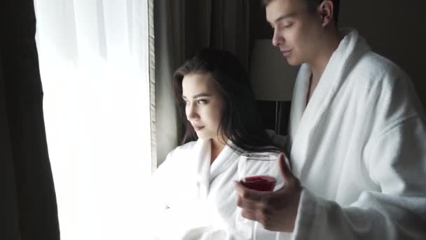 Loving couple in white coats looks at landscape outside the window, the guy brings the girl a glass of red wine stock footage video - Materiał filmowy, wideo