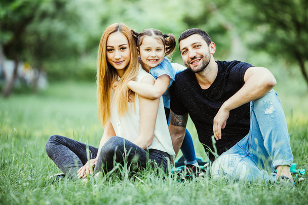 Young happy family in summer park. Father and mother spend time in summer park with their daughter while she hug them. - Photo, Image