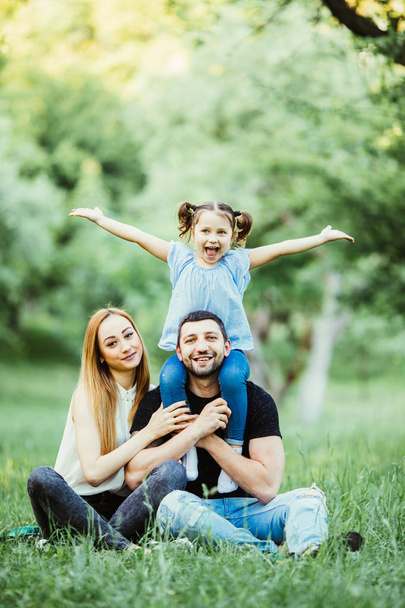 Young happy family of three having fun together outdoor. Pretty little daughter on her father piggyback smiling and have fun. - Photo, Image