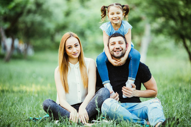 Happy mother, father and daughter in the park. Beauty nature scene with family outdoor lifestyle. Happy daughter on father back smile. - Photo, Image