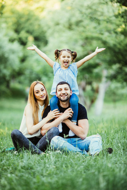 Young happy family of three having fun together outdoor. Pretty little daughter on her father back with happy raised hands. Parents and girl look happy and smile. - Photo, Image