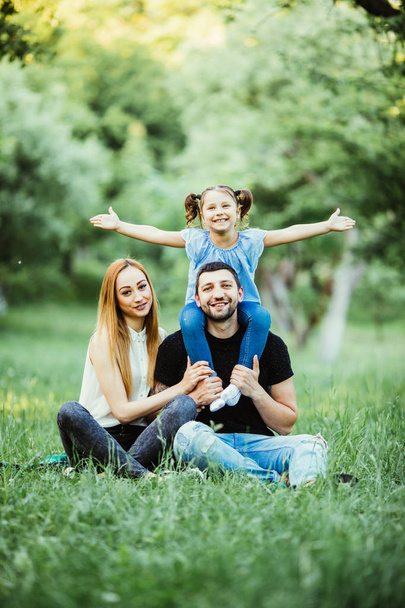 Young happy family of three having fun together outdoor. Pretty little daughter on her father back with happy raised hands. Parents and girl look happy and smile. - Photo, Image