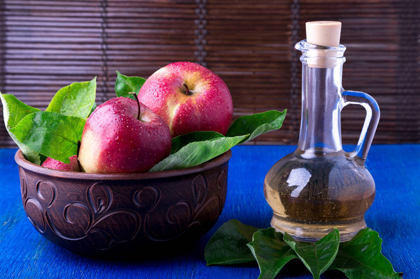 Vinagre de sidra de manzana en botella de vidrio sobre fondo azul. Manzanas rojas en tazón marrón
. - Foto, imagen