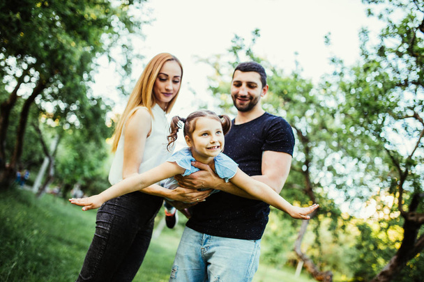 Happy joyful young family father, mother and little daughter having fun outdoors, playing together in summer park. Daughter raided hands try to fly in parents hands - Φωτογραφία, εικόνα
