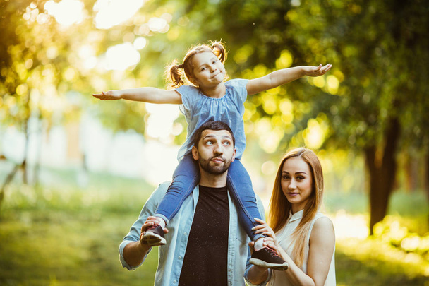 Ευτυχισμένη οικογένεια παίζει στην φύση αργά απογευματινού ήλιου, το φθινόπωρο, καλοκαίρι. Μητέρα, πατέρας και κόρη παίζει στο γρασίδι στο πάρκο, γελώντας και χαμογελαστά, διασκέδαση, χαρά. - Φωτογραφία, εικόνα