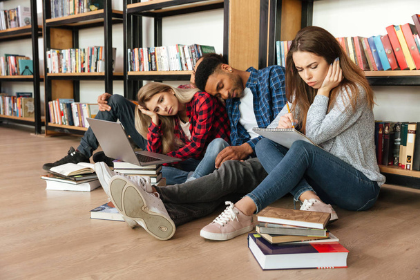 Молодые уставшие студенты сидят в библиотеке на полу
 - Фото, изображение