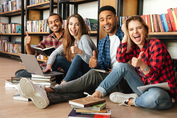 Smiling students sitting in library on floor using laptop - Φωτογραφία, εικόνα