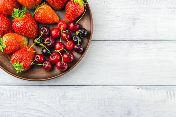 Ernte frischer Bio-Beeren, Erdbeeren und Kirschen in braunem Tonteller auf weißem Holzdielen-Hintergrund, Draufsicht. - Foto, Bild