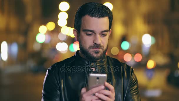 笑みを浮かべてラテン男は、夜背景で街の明かり、スマート フォンで sms を送信します。 - 映像、動画