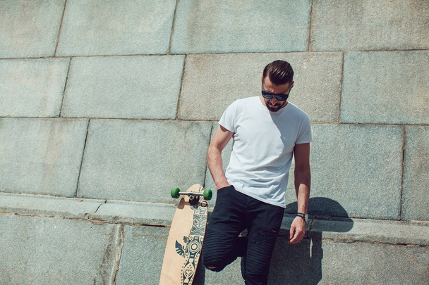 Stylish man in sunglasses and with a beard stands on wall background with a long board. T-shirt mock up. - Foto, Imagen