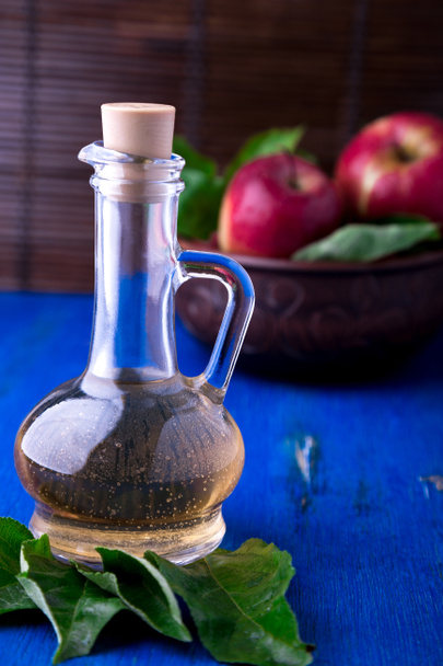 Apple cider vinegar in glass bottle on blue background. Red apples in brown bowl. - Photo, Image