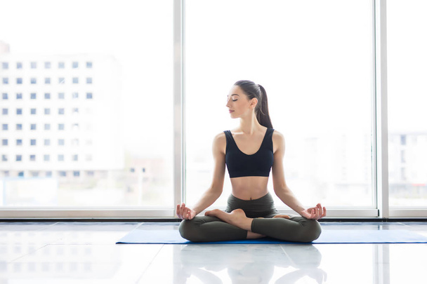 Поза медитационного лотоса. Молодая привлекательная женщина практикующая йогу, сидящая в упражнениях, тренирующаяся, носящая спортивную одежду у окна этажа с видом на город
 - Фото, изображение