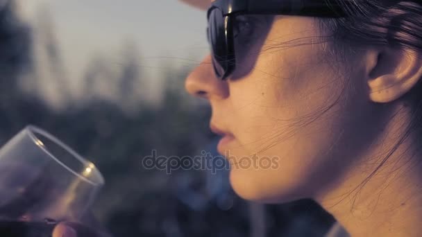 Młoda piękna dziewczyna siedzi w profilu i podziwiając zachód słońca, picie czerwonego wina, lekkie wiatr podnoszenia jej włosy. Z bliska. slowmotion. HD. 1920 x 1080 - Materiał filmowy, wideo
