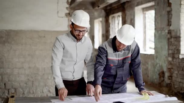 ein erwachsener Ingenieur erklärt dem Arbeiter einen neuen Bauplan, der vom Architekten entworfen wurde, die Menschen befinden sich in einem verlassenen Gebäude - Filmmaterial, Video