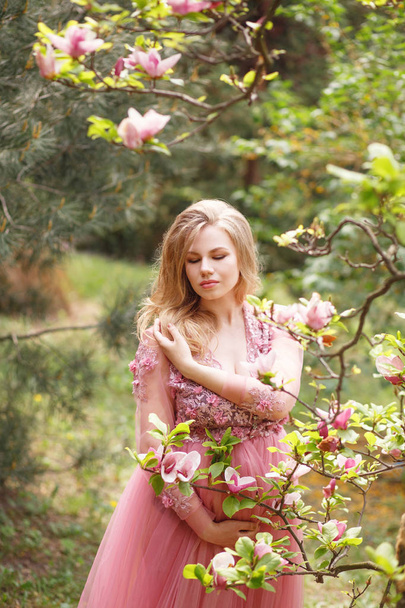Młoda piękna dziewczyna w długiej sukni dotyka jej ciąży brzuch w pobliżu kwitnący magnolia Park - Zdjęcie, obraz