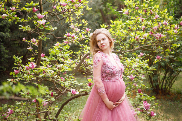 Прекрасна жінка очікує дитину на красивому платті на фоні цвітіння магнолії та зелені
 - Фото, зображення