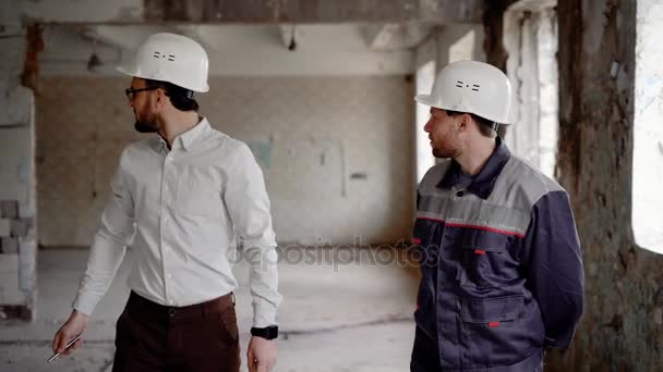 Un hombre de negocios con un casco en la cabeza y un superintendente de construcción con uniformes de protección están considerando la estructura para la futura reconstrucción
 - Imágenes, Vídeo