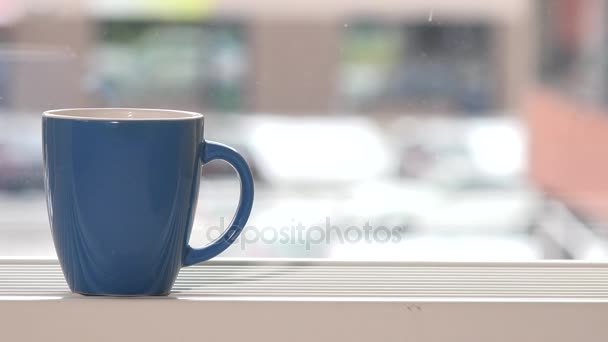 Blaue Tasse Heißgetränk steht auf Fensterbank und Schneeflocken fallen - Filmmaterial, Video