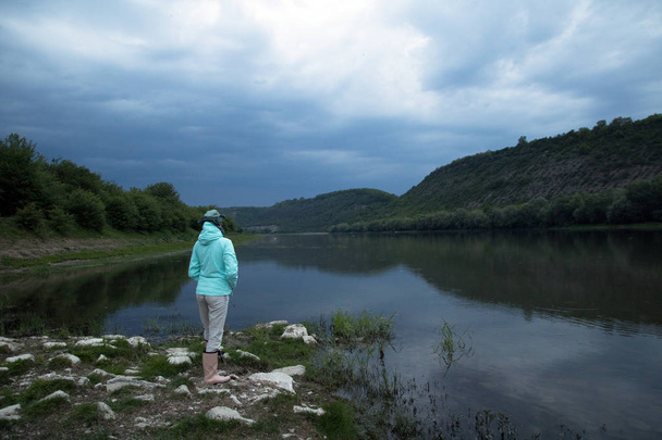 女性は川の近くに立って、遠くを見ます。夕暮れ時の青い嵐雲 - 写真・画像