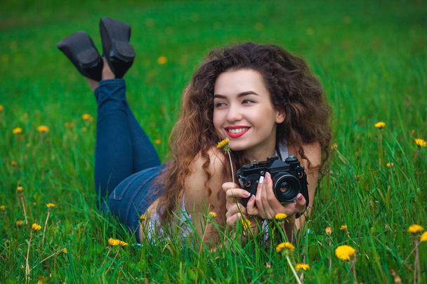 Mooi meisje-fotograaf met krullend haar bezit een camera en liggen op het gras met bloeiende paardebloemen in het voorjaar buiten in het park. - Foto, afbeelding