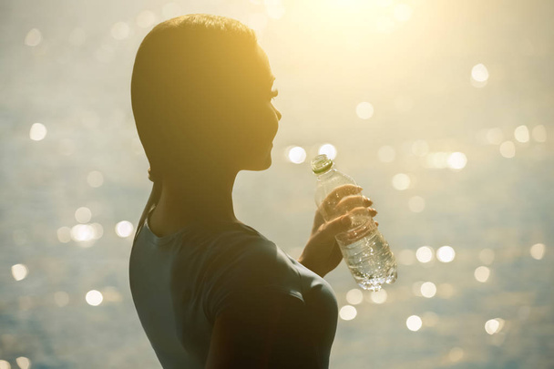 Σιλουέτα του μια νεαρή αθλήτρια σε αθλητικές φόρμες πόσιμο νερό από ένα μπουκάλι  - Φωτογραφία, εικόνα