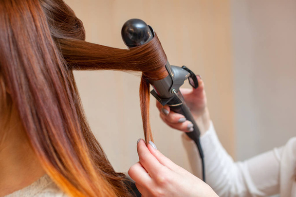 Salon fryzjerski sprawia, że fryzura dziewczyna z długimi rudymi włosami w salonie kosmetycznym. Stwórz Loki z lokówki. - Zdjęcie, obraz