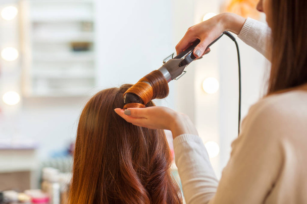 Salon fryzjerski sprawia, że fryzura dziewczyna z długimi rudymi włosami w salonie kosmetycznym. Stwórz Loki z lokówki.  - Zdjęcie, obraz