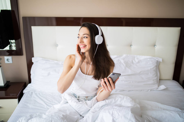 Piękna dziewczyna w słuchawkach jest słuchanie muzyki za pomocą smartfona, patrząc na kamery i uśmiechając się leżąc na łóżku w domu - Zdjęcie, obraz
