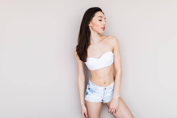 beautiful young woman with a sports figure in denim shorts and a black bra standing on a white background - Foto, Imagem