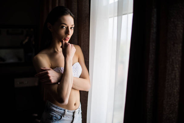 Beautiful woman near curtains of window and looking at the camera - Фото, изображение