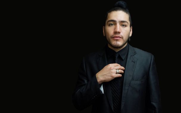 Portrét mladého muže Latinské nosí náušnice černý oblek, černou košili a černou kravatu na černém pozadí s rukou na kravatu pohledu zepředu - Fotografie, Obrázek