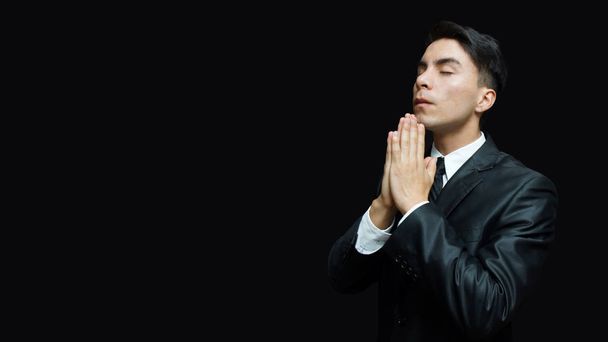 Latino jongeman in zwarte pak, wit overhemd, zwarte stropdas met zijn handen in een gebed standpunt - Foto, afbeelding