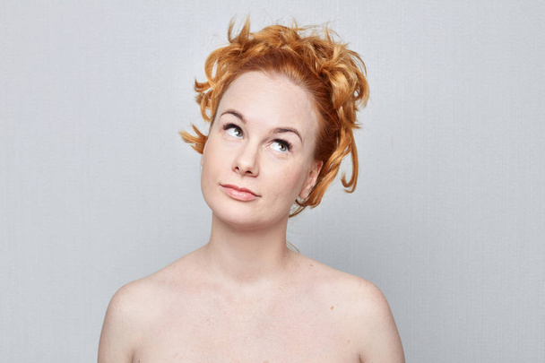 Human face expressions, emotions and feelings.Nude studio portrait of beautiful young Caucasian redhead freckled woman looking playfully sideward demonstrating something on grey wall with copy advertising space. - Fotografie, Obrázek