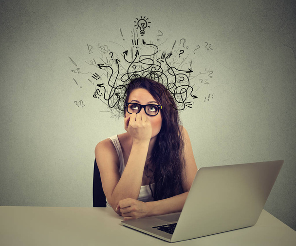 woman with thoughtful expression sitting at a desk with laptop with arrows and symbols coming out of head - Foto, afbeelding