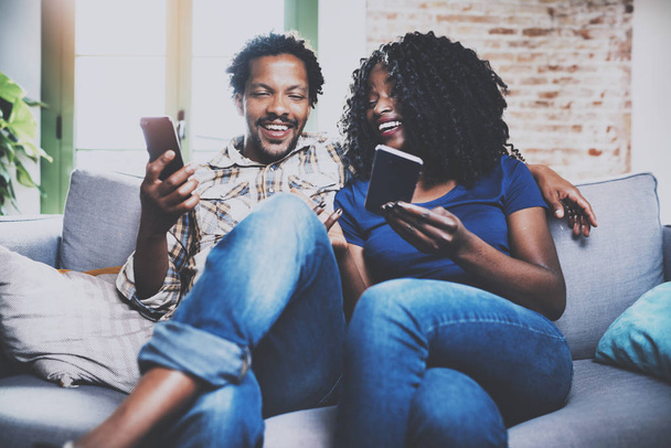 Sonriente pareja afroamericana relajándose juntos en el refresco.Joven hombre negro y su novia usando dispositivos móviles mientras descansan en casa en la sala de estar. Fondo horizontal, borroso
. - Foto, imagen