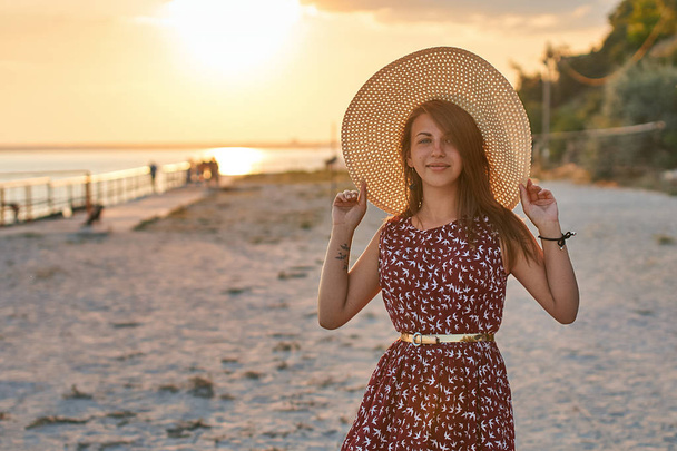 Fashion portrait of a young beautiful girl in a dress with bird pattern holding a straw beach hat and being happy in the rays of sunset sunlight at the summer seaside - Foto, afbeelding