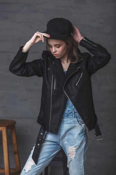 Young teenage girl wearing leather jacket jeans and cap, studio portrait over dark background - 写真・画像