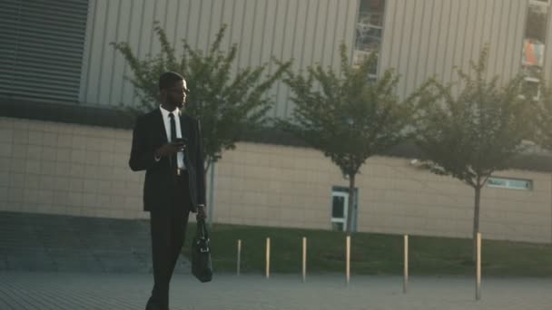 Attractive african american businessman walking near office center holding smartphone, reading messages, surfing internet. - Felvétel, videó