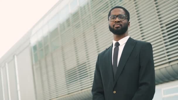 Ritratto ravvicinato di attraente uomo d'affari afro-americano in abito nero sorridente alla telecamera otdoors vicino all'edificio del centro ufficio
. - Filmati, video