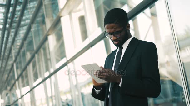 Happy african american businessman using tablet device while standing near big office windows outdoors. Man scrolling pages, tapping on touchscreen, checking financial charts. close up - Felvétel, videó