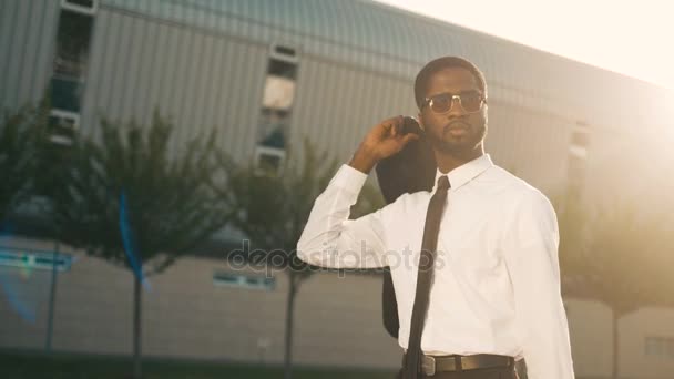 Attractive black african american businessman walking near office center holding jacket over his shoulder and smiling. Man going home after work. Sun rays. Close up - Πλάνα, βίντεο