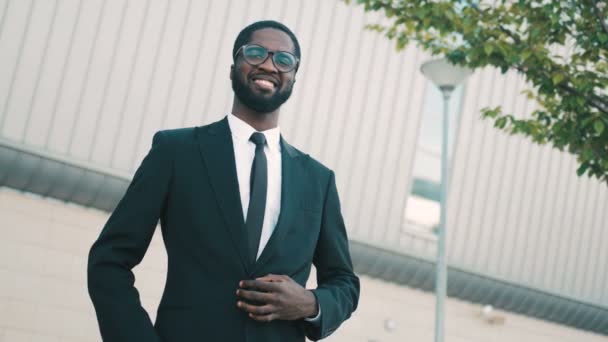 Happy attractive african american businessman standing near office center building in fashion suit, looking straight at the camera and smiling. Portrait shot - Filmati, video