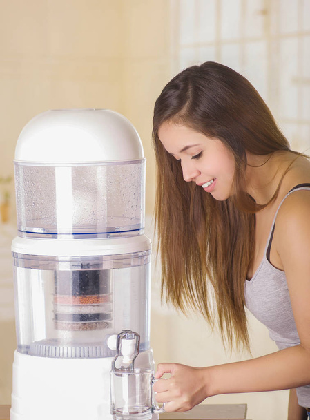 Beautiful smiling woman filling a glass of water, with a filter system of water purifier on a kitchen background - Photo, Image