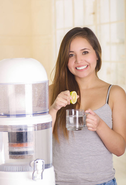 Beautiful smiling woman holding a glass of water behind of a filter system of water purifier on a kitchen background - 写真・画像